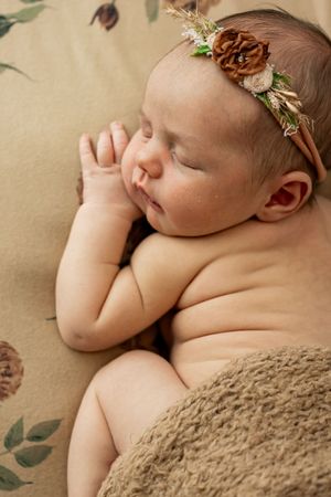 Peaceful sleeping newborn wearing pine cone and flower headband on tan floral fabric.