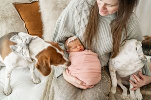 A swaddled newborn rests on a lap in a cozy sweater while two dogs cuddle close on either side.