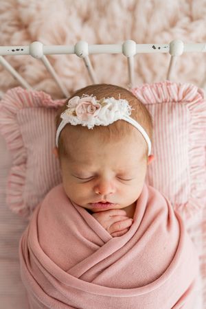 Sleeping newborn wrapped in soft pink swaddle with delicate white floral headband against light background.