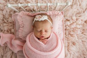 Sleeping newborn baby wrapped in soft pink knit blanket with delicate lace headband against textured blush backdrop.