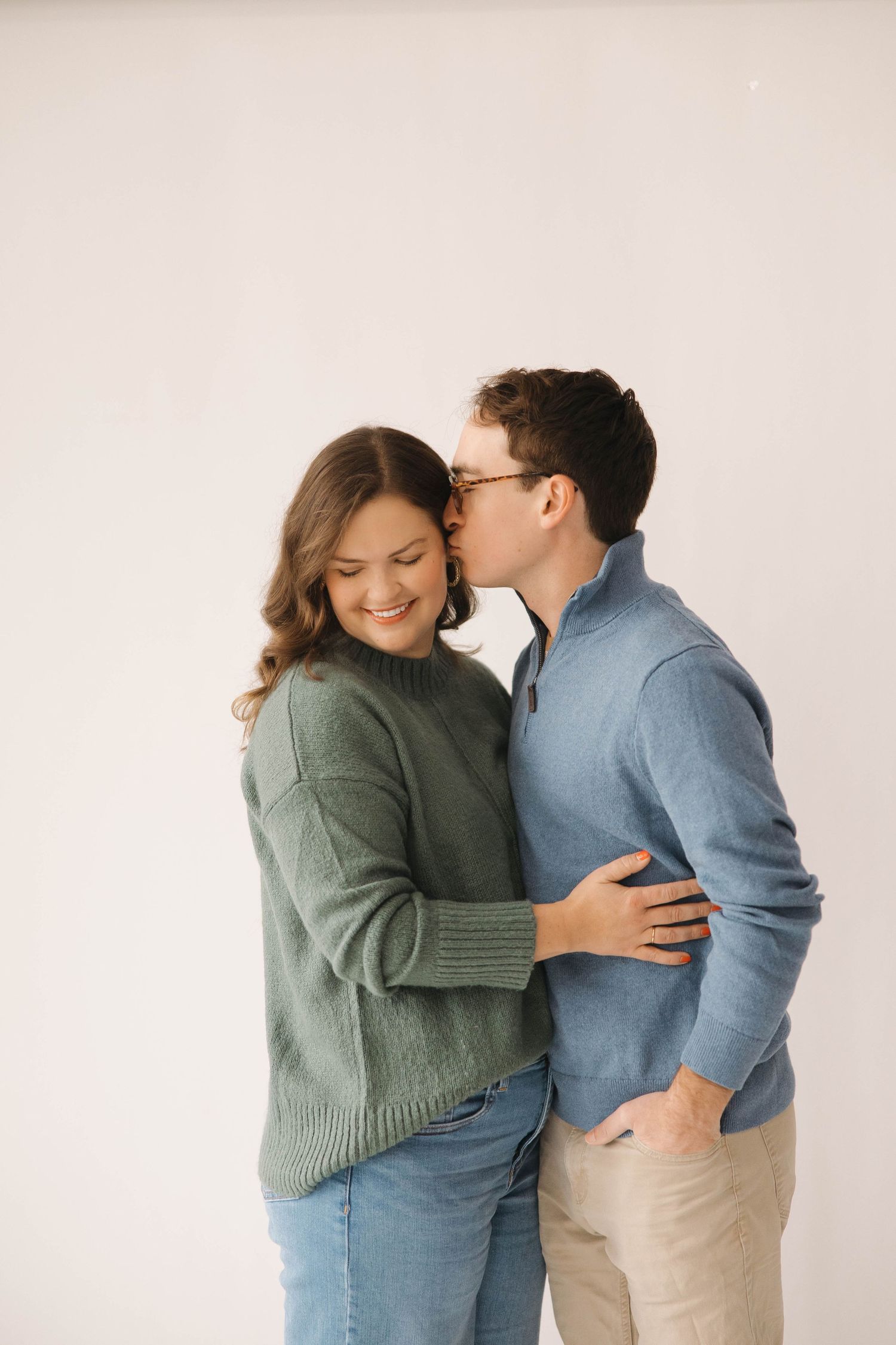 A couple in casual sweaters share an intimate moment while embracing against a light background.