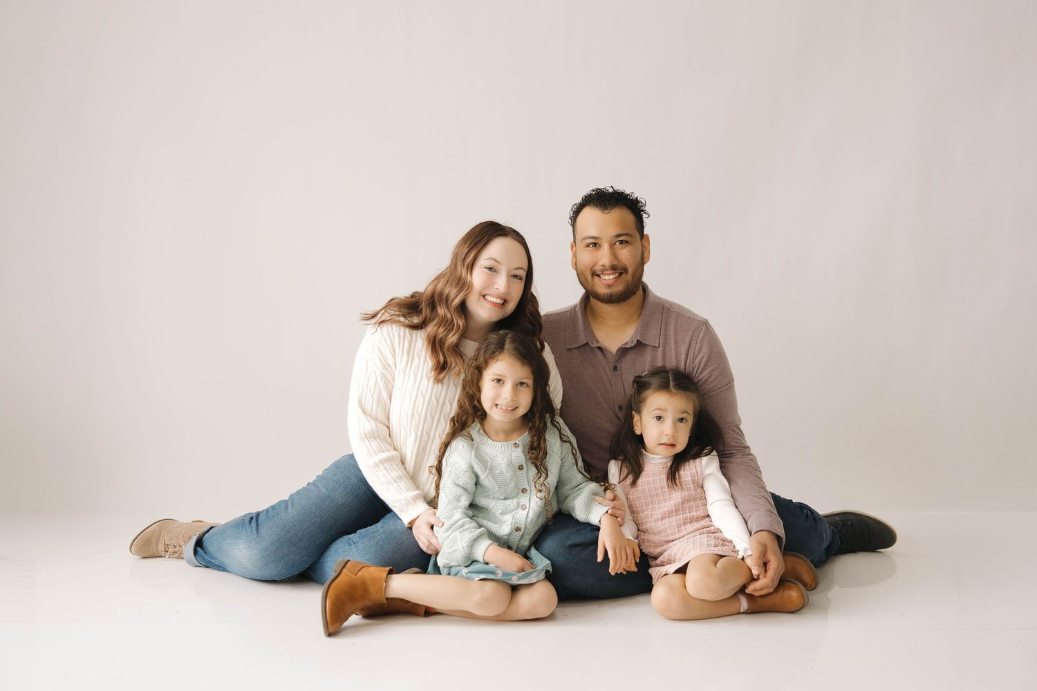 A family of four sits together on a white backdrop wearing casual clothing in a studio portrait setting.