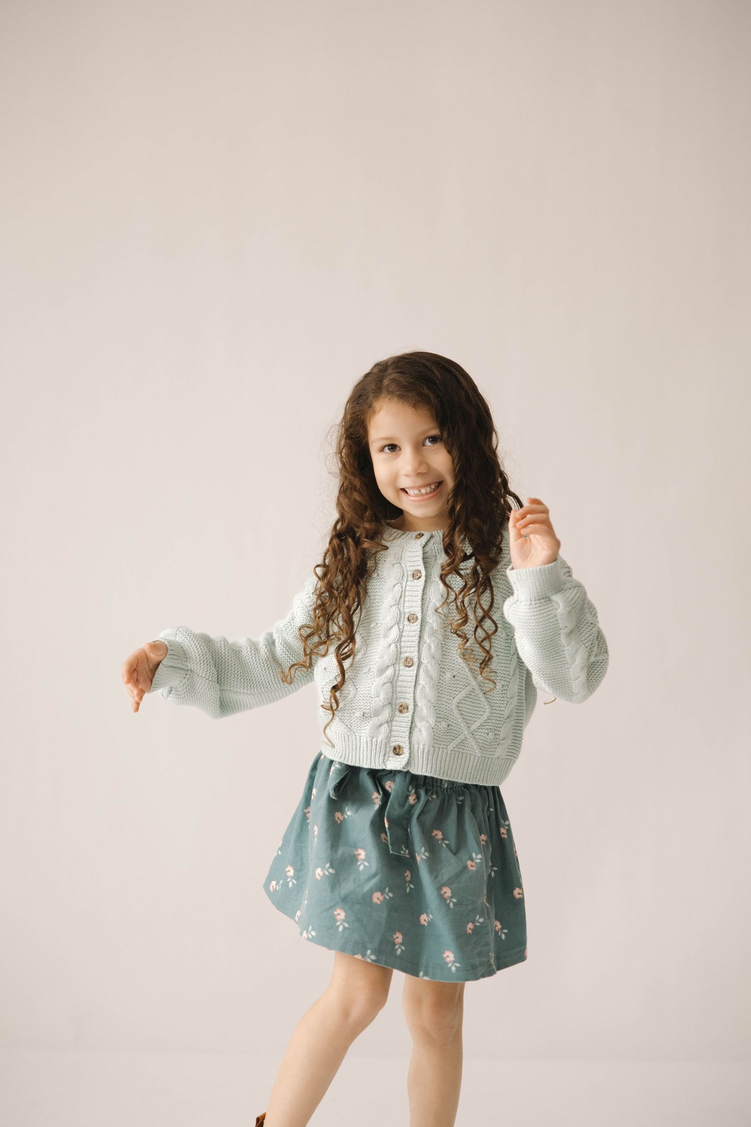 A young child in a mint cardigan and floral dress dances and twirls in a bright studio setting.