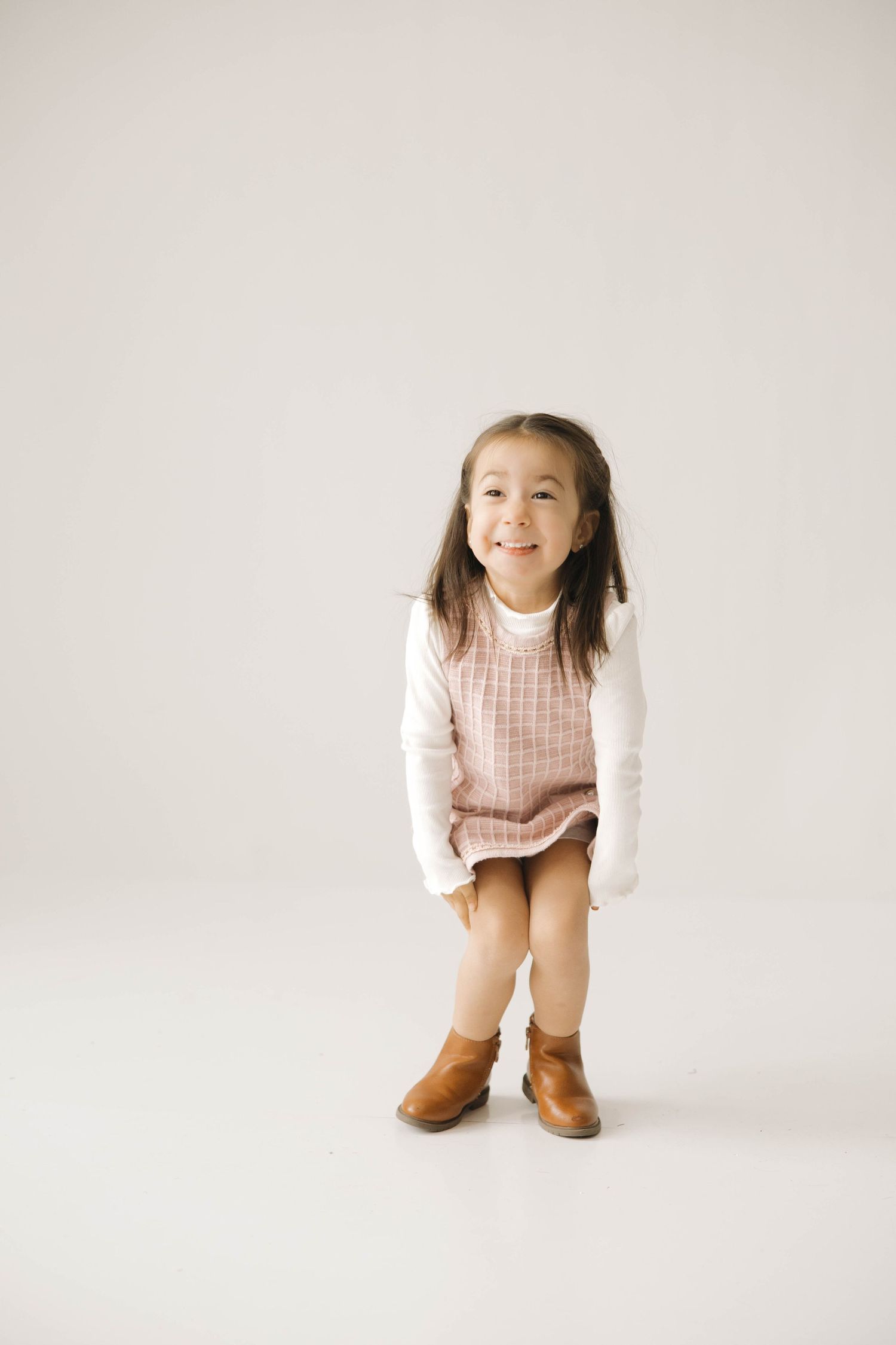 Child in a pink jumper dress and white long sleeve shirt poses playfully against white background.