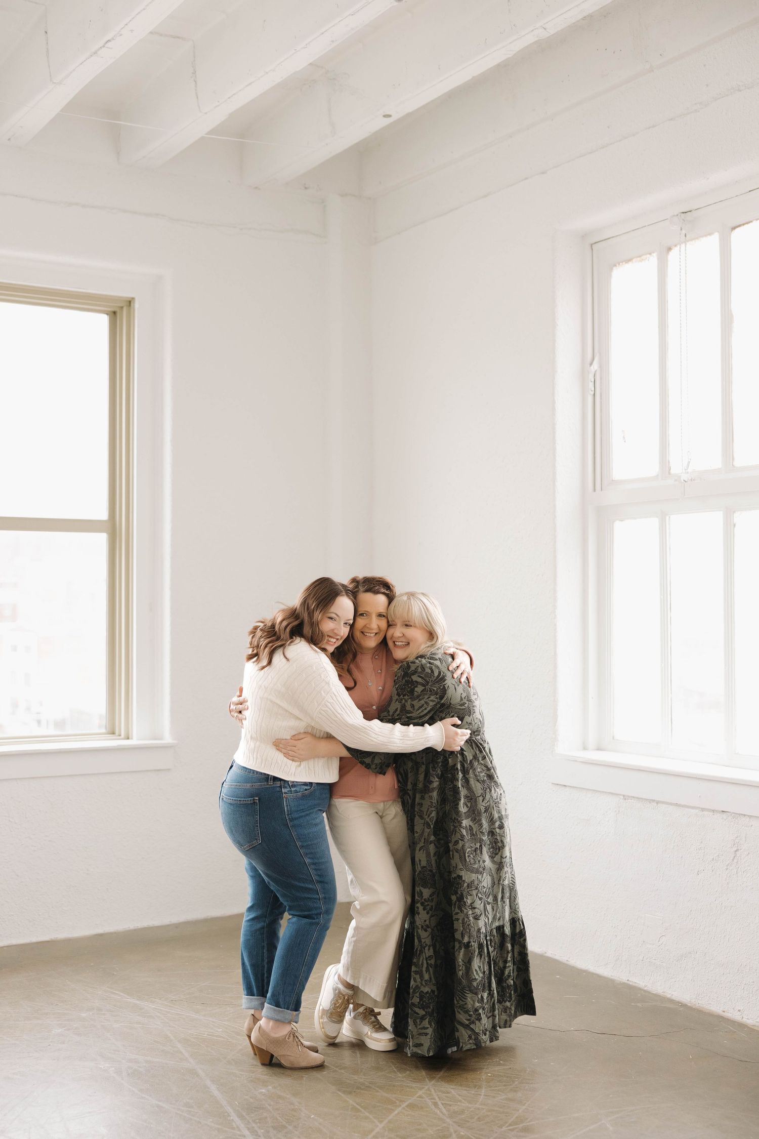 Three people embrace in a tender moment against a white wall with bright windows.