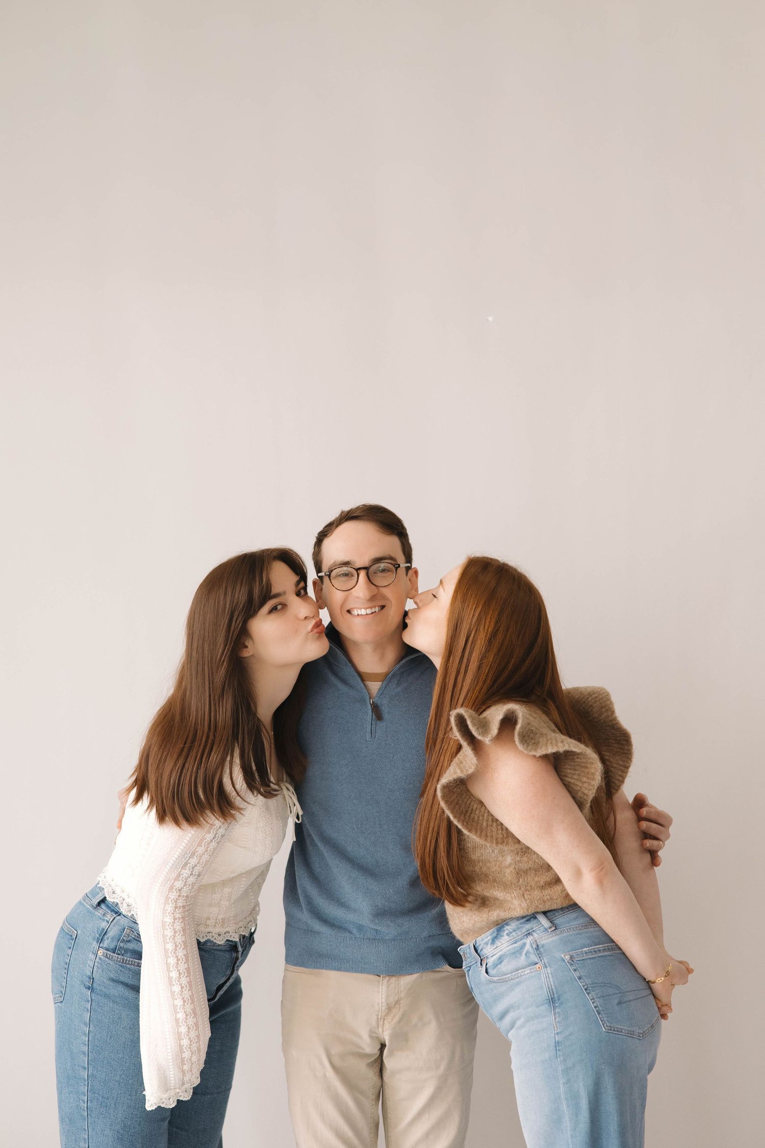Three people pose together against white wall while wearing casual denim and sweaters in progressive kissing sequence.