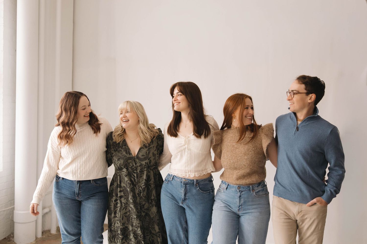 A group of people wearing casual denim and neutral tones stand together against a white wall.