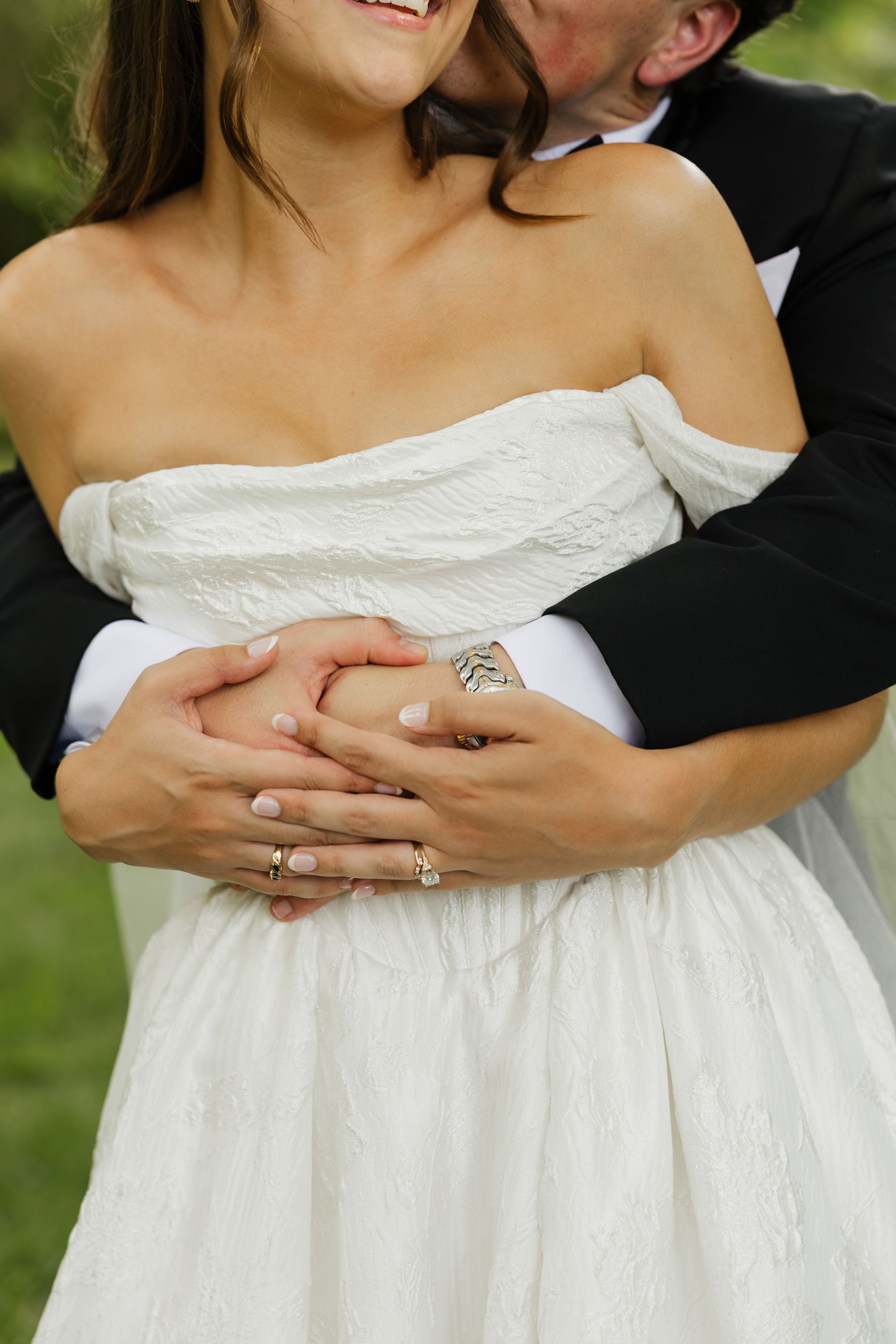 Groom in black suit embracing bride in white off-shoulder dress, hands clasped together showing wedding rings outdoors.
