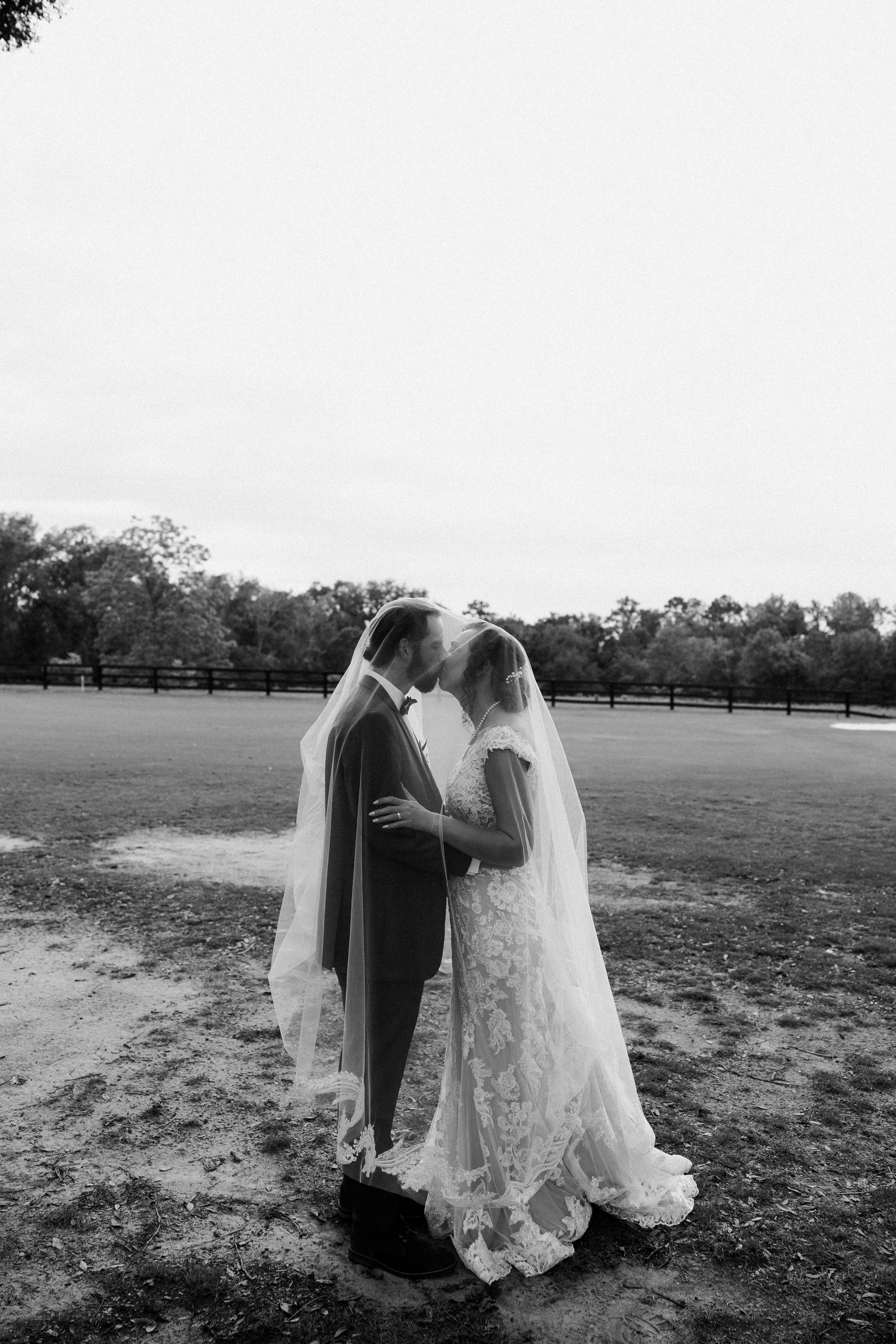 Black and white photo of a bride and groom sharing a kiss under her veil in an open field.