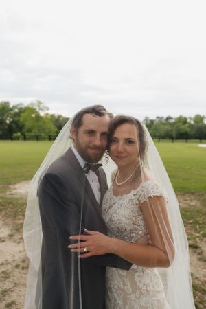 Bride and groom pose together under a flowing veil outdoors, with the bride in lace dress and pearl necklace.