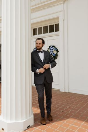 Groom in black tuxedo with bow tie adjusting jacket beside white column at wedding venue.