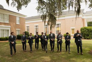 Groomsmen in dark suits walking together on green lawn outside elegant brick building with Spanish moss trees.