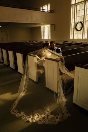 Bride and groom sit intimately in church pews, her lace cathedral veil cascading dramatically across the aisle in golden light.