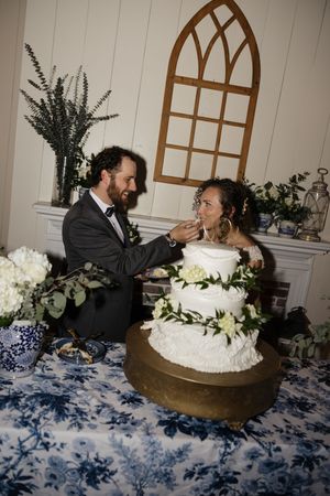 Bride and groom cutting white three-tier wedding cake decorated with greenery at reception.