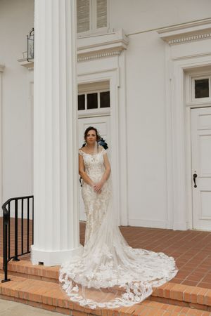 Bride in lace mermaid wedding gown with long train standing by white column at elegant venue entrance.