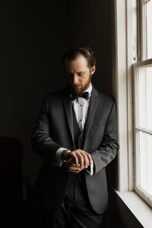 Groom in gray tuxedo and bow tie adjusts cufflinks by window in moody natural light.