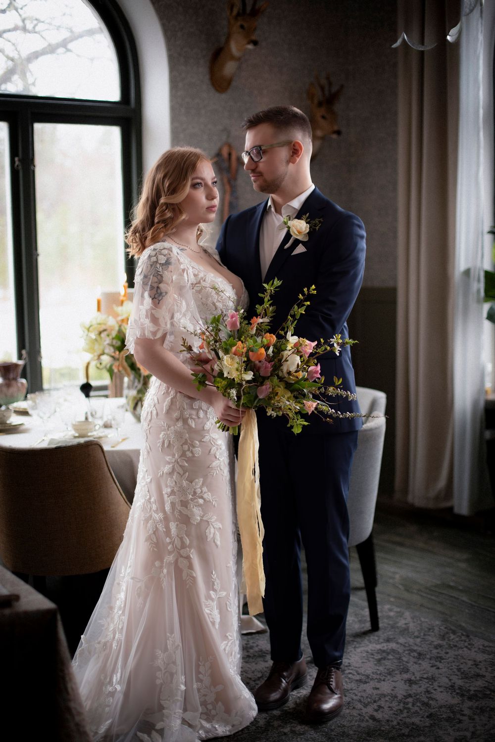 bröllopsfotografering inomhus. bruden håller i en bukett och brudgummen håller om bruden. mannen  har en blå kostym med en gul ros i fickan.