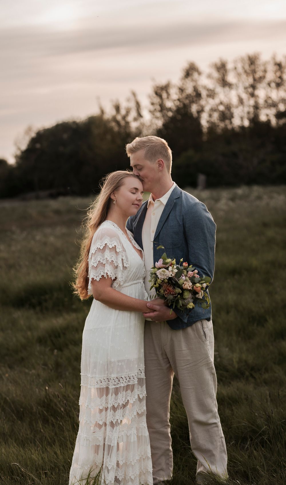 bröllopsfotografering på en äng. Bruden bär en vit lång klänning och håller en bukett. Brudgummen  en vit skjorta, blå kavaj och beiga byxor