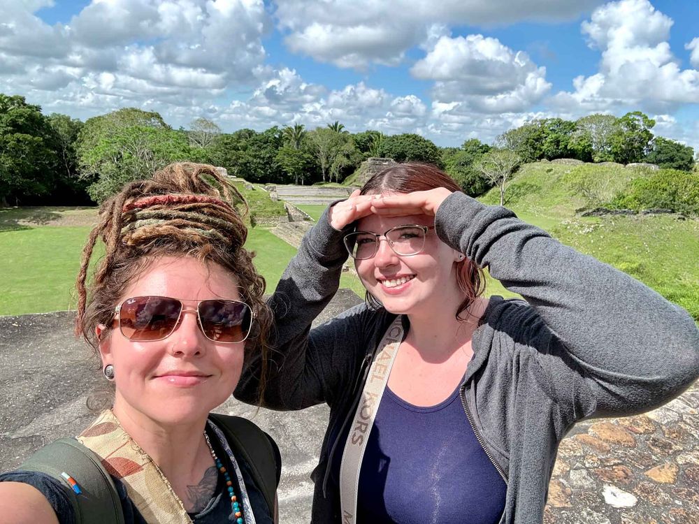 Two people take a selfie outdoors on a sunny day with puffy white clouds and green hills in the background.