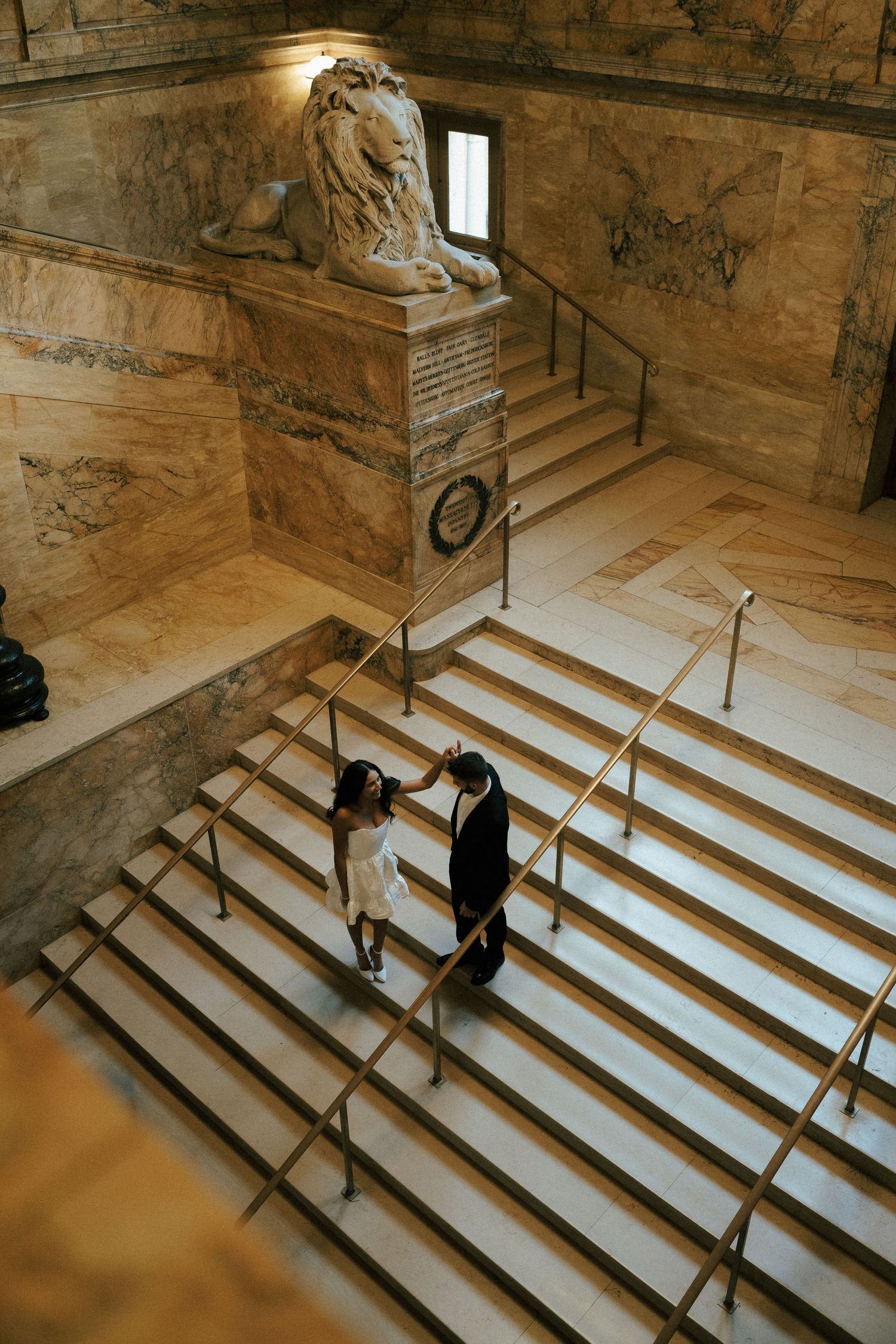 Two people stand on a grand marble staircase in a historic building with ornate lion sculpture and architectural details.