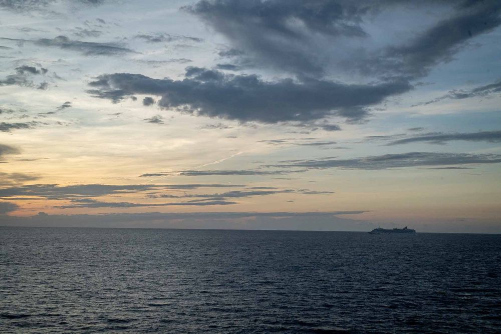 Dramatic sunset over calm ocean waters with dark clouds and distant ships on the horizon.