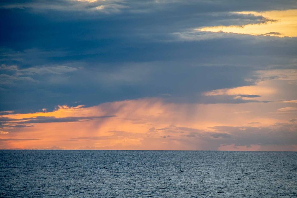 Dramatic sunset sky with orange rays breaking through dark clouds over a calm ocean horizon.