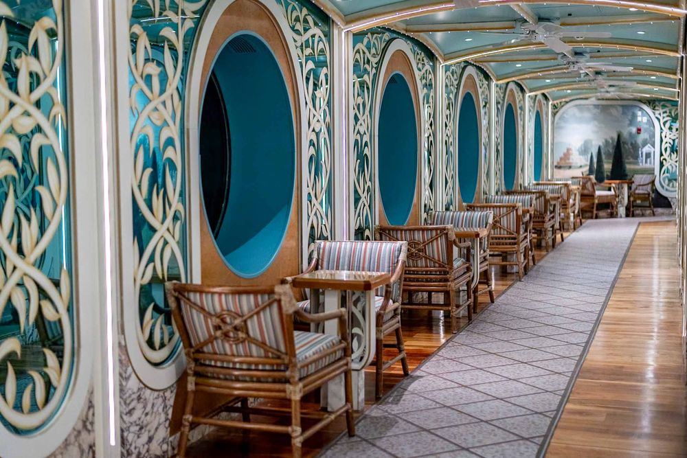 Long ornate hallway with turquoise and gold decor features wooden benches and circular windows along the wall.