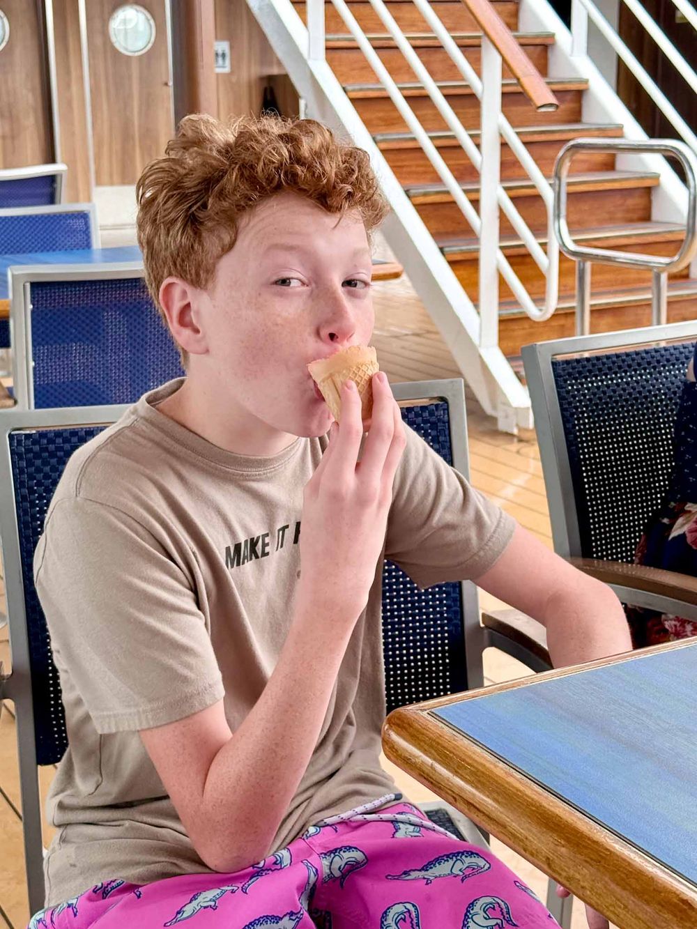 Student in grey t-shirt sits at desk in classroom making shushing gesture with finger to lips.