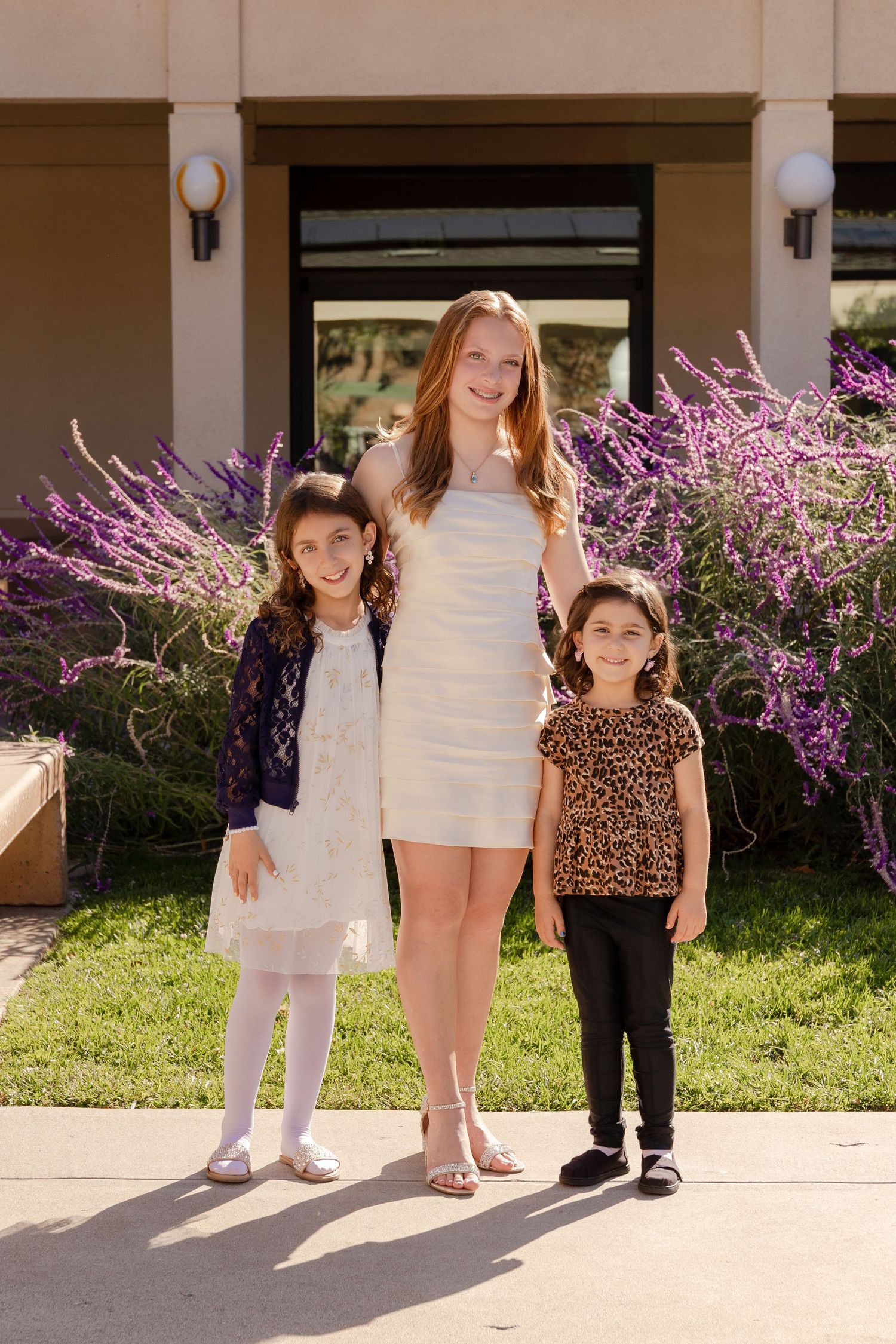 A group of children and adults stand together outside a building with blooming pink flowers in the background.