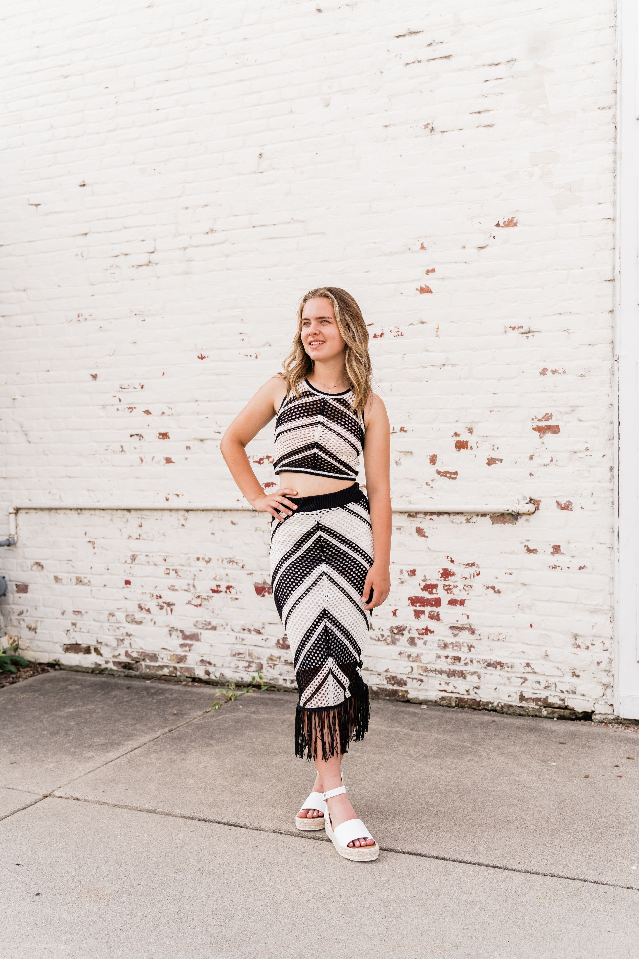 A striped black and white dress is styled with white sandals against a distressed brick wall.