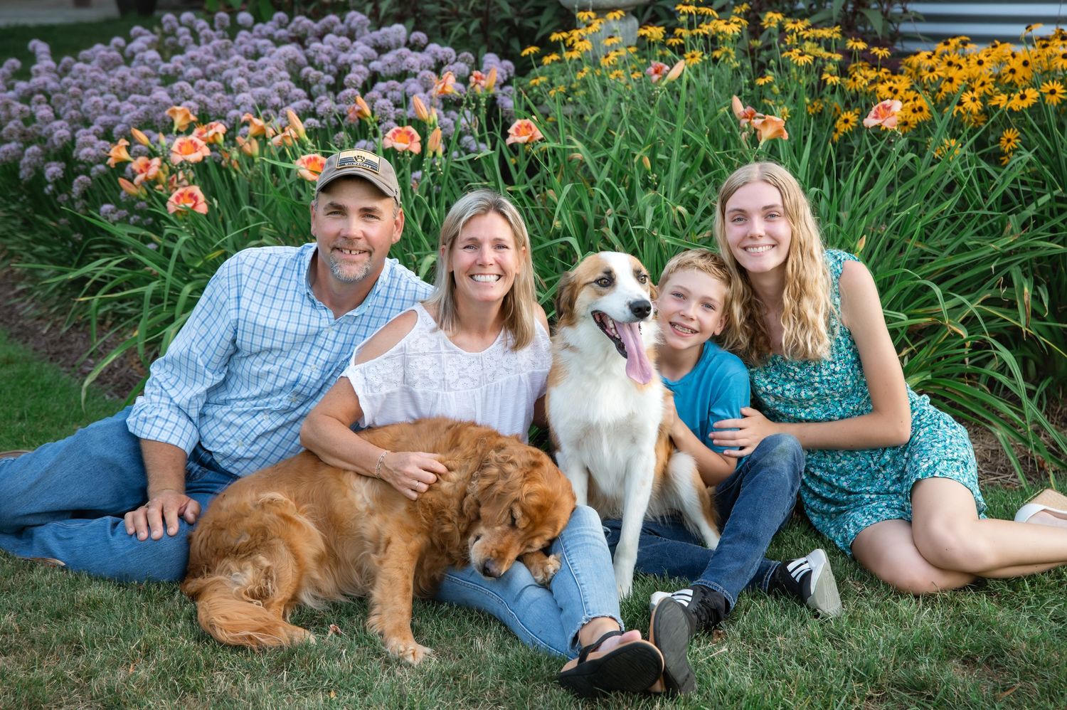 A group relaxes on grass with two dogs in front of a colorful garden with purple and orange flowers blooming.