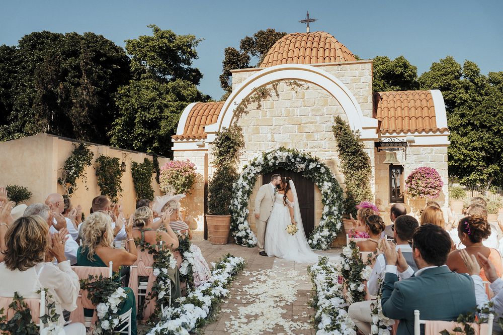 Outdoor wedding ceremony with white floral arch and guests seated on wooden chairs under blue summer sky.