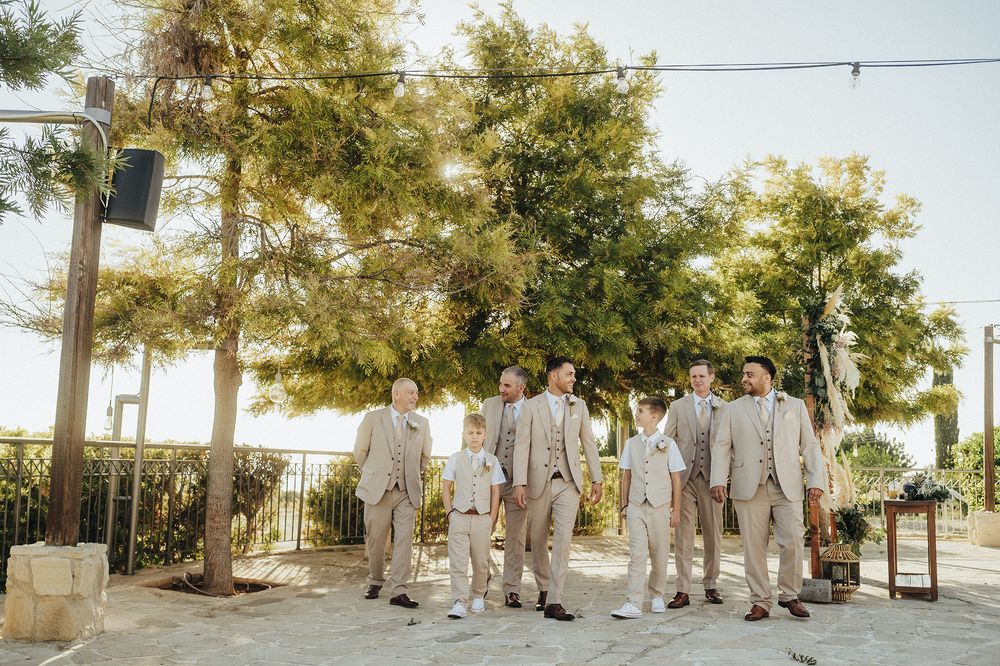 A wedding party in light beige suits walks in a line outdoors against a backdrop of trees and sunlight.