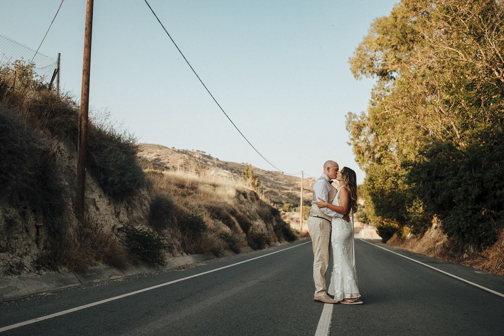 A couple stands on a winding mountain road surrounded by golden trees and rocky cliffs at sunset.