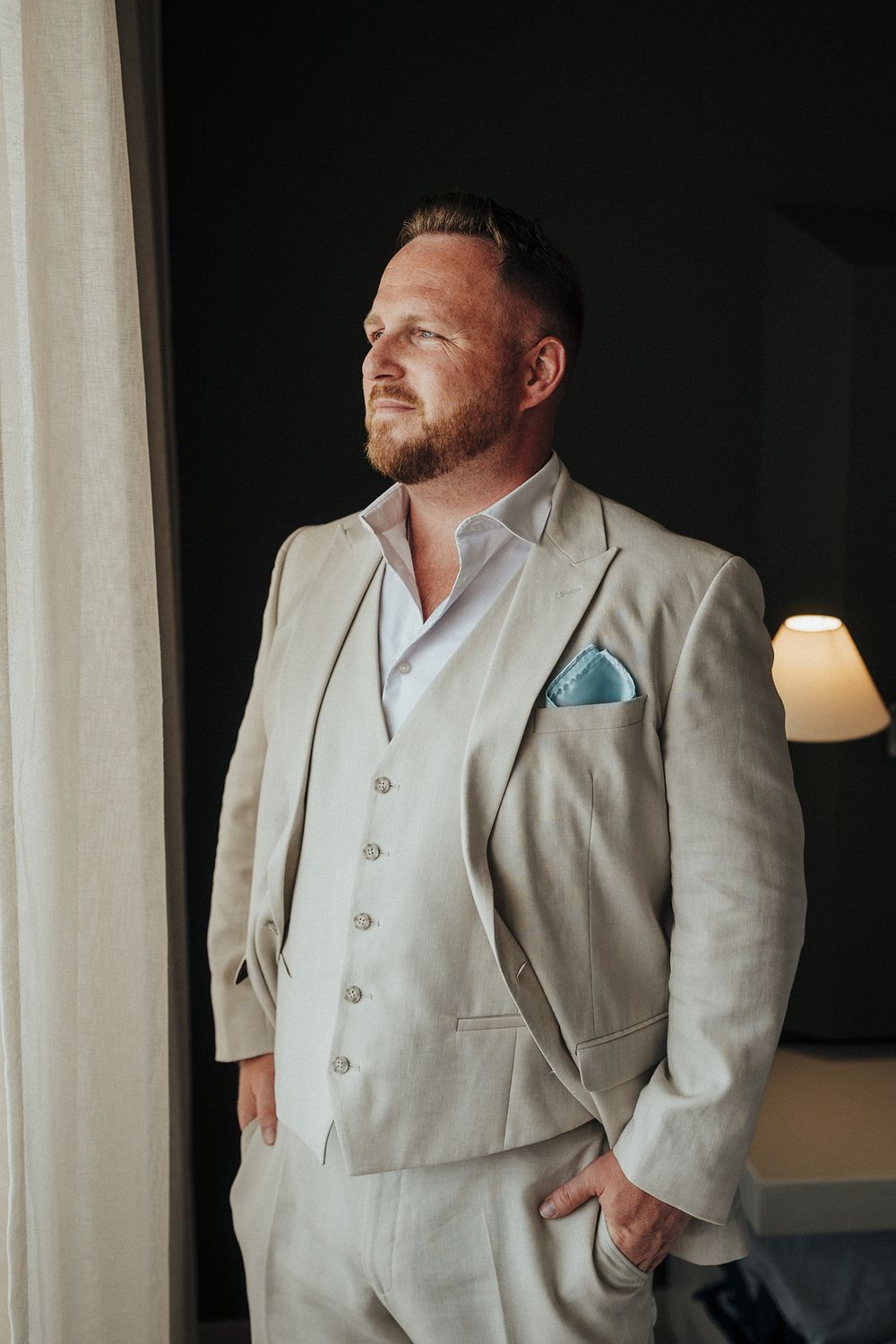 A person in a light beige suit poses thoughtfully near a window in a dimly lit room with white curtains.
