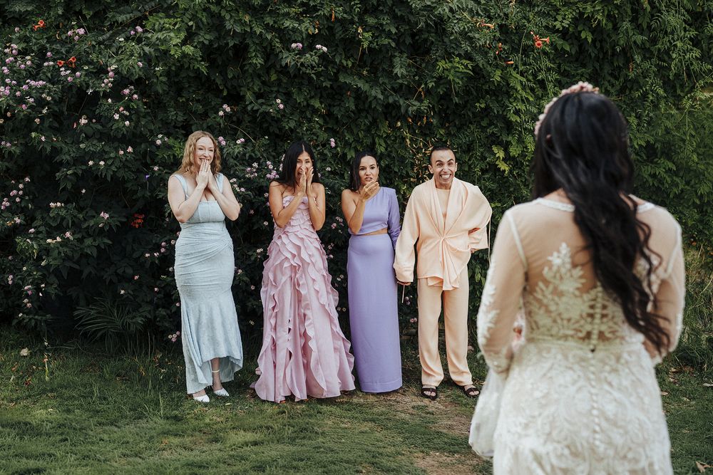 Wedding party members react with joy as they watch a bride in a lace dress during an outdoor garden celebration.