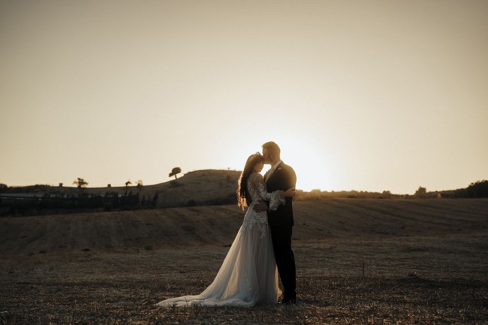 A couple in wedding attire embraces on a hilltop at sunset with a dramatic sky in the background.