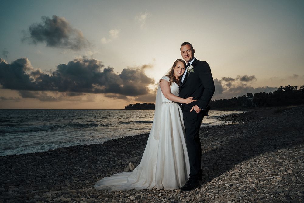 A romantic beach wedding photo at sunset with a couple embracing on the rocky shoreline.