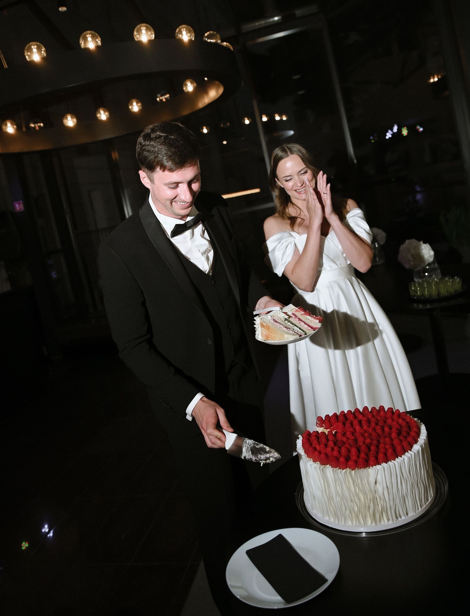 A couple cuts into a red and white wedding cake with dramatic lighting at their reception celebration.