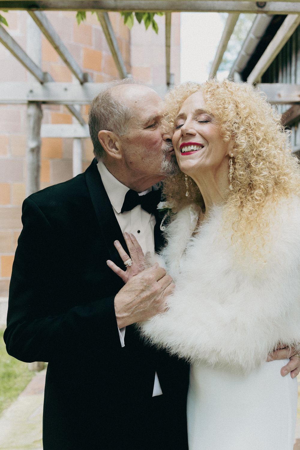 A couple shares a tender moment in formal attire, with the woman wearing a white dress and fur wrap while laughing joyfully.