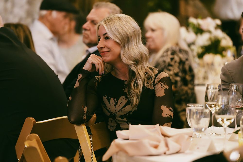 Woman in black lace dress smiles while seated at elegant dinner table with wine glasses and place settings.