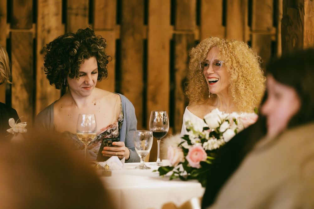 Wedding guests laugh and socialize around a rustic wooden table during an intimate dinner reception.