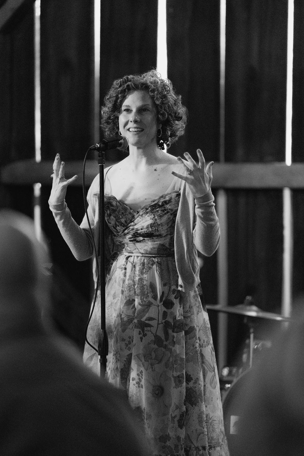 A performer in a floral dress gestures expressively on stage with dramatic lighting and vertical bars in the background.