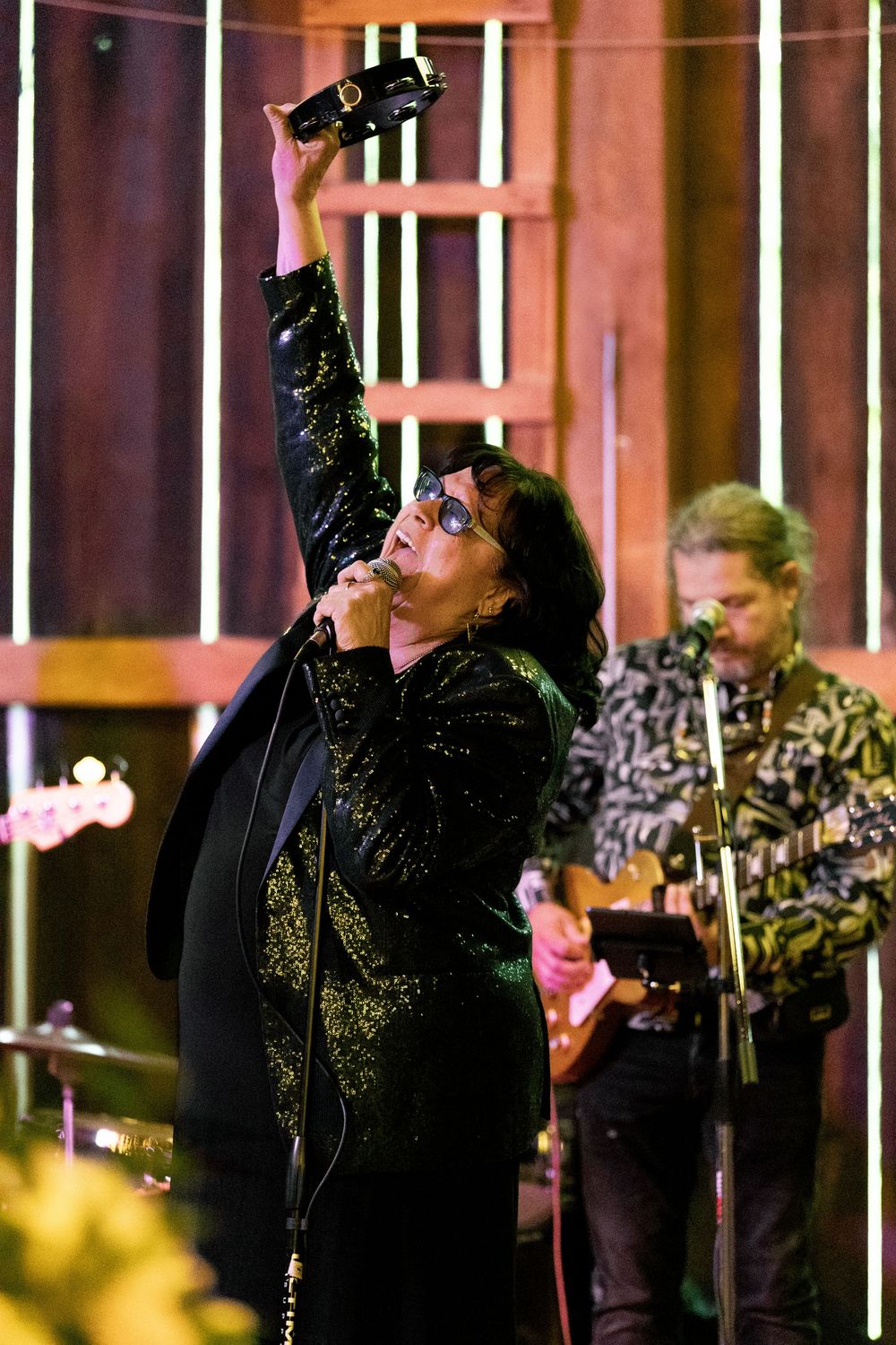 A performer in black sequined attire raises their arms during an energetic stage performance with musicians behind them.