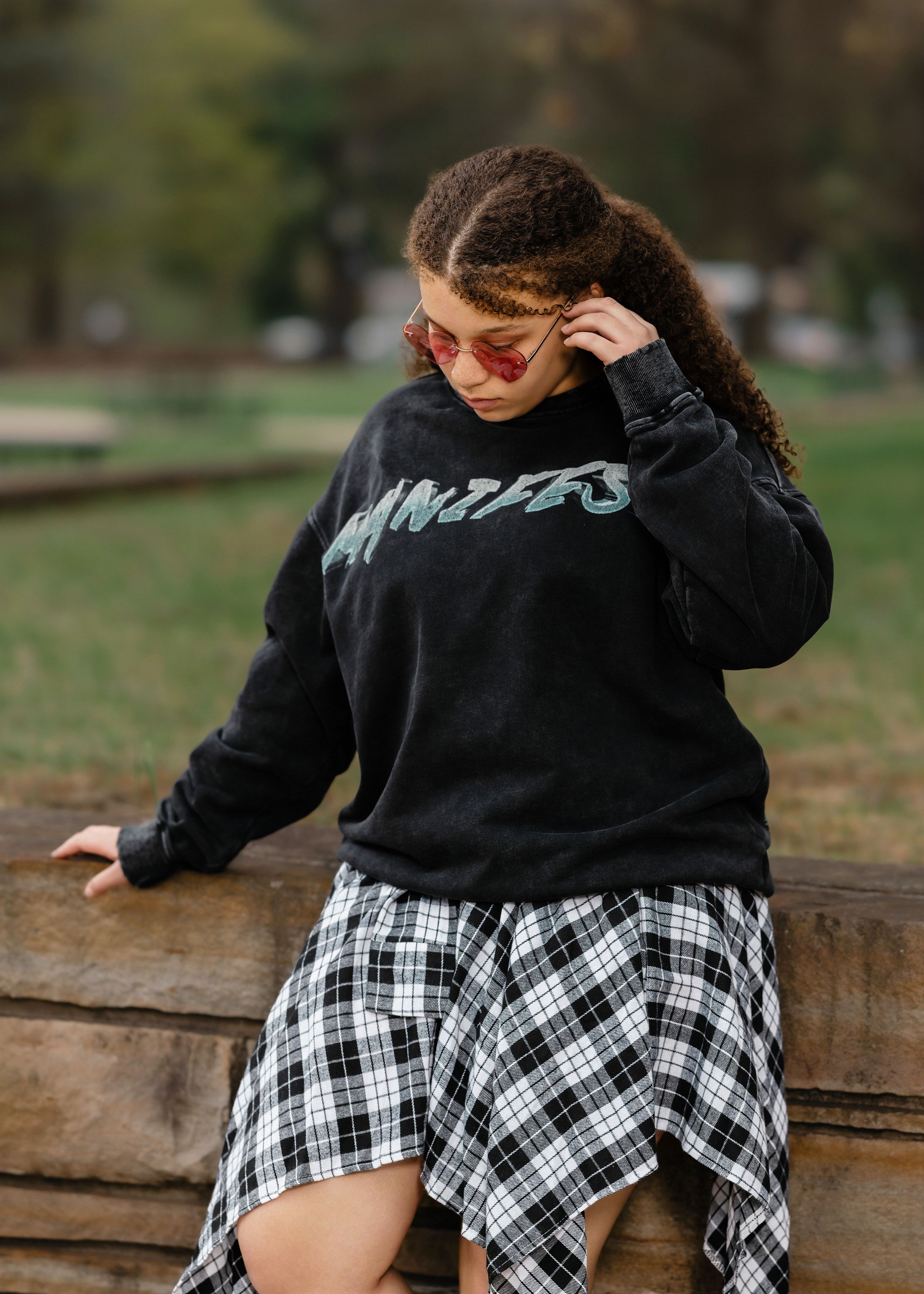 Person in black sweater and plaid skirt sitting on stone wall outdoors.