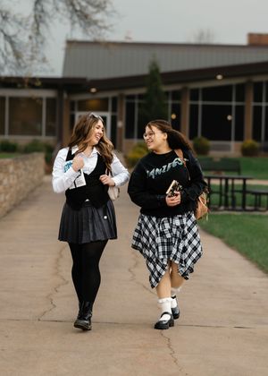 Two friends in coordinating black and plaid outfits walk and laugh together on a campus sidewalk.
