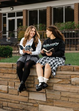 Friends in school uniforms sit and chat on a stone wall outside a brick building.