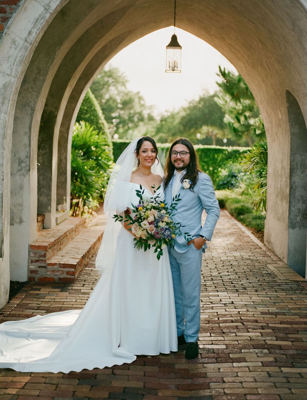 Wedding party poses in a beautiful brick archway corridor with natural lighting and elegant floral bouquets.