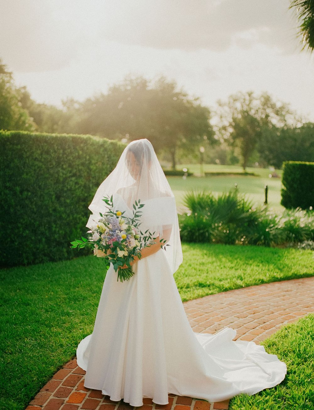 A white wedding dress with a flowing train and veil is photographed in a sunlit garden with brick pathways.