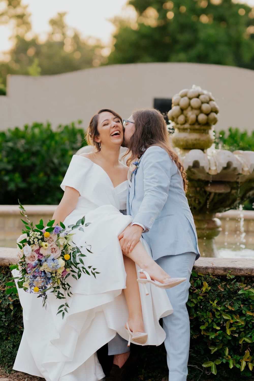A joyful wedding couple shares a romantic moment in front of a garden fountain during golden hour.